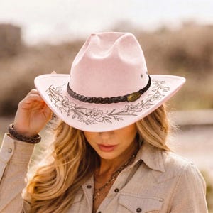 Pode incluir: Um chap&eacute;u de cowboy rosa claro com uma faixa tran&ccedil;ada castanho-escura e um detalhe oval met&aacute;lico. A aba &eacute; decorada com uma ilustra&ccedil;&atilde;o de linhas pretas de motivos florais, incluindo narcisos e caules com folhas ao redor da copa.