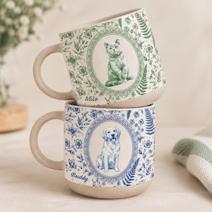 May include: Two ceramic mugs with a speckled beige base. The top mug features a green floral pattern, an oval frame with a cat, and the name Milo. The bottom mug features a blue floral pattern, an oval frame with a dog, and the name Buddy.