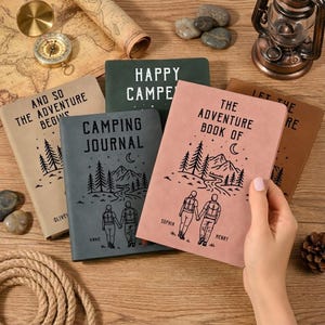 May include: A collection of adventure-themed journals in various colors, including pink, green, and tan. Each journal features unique text and illustrations of mountains, trees, and people. The journals are personalized with names and phrases like "Camping Journal."
