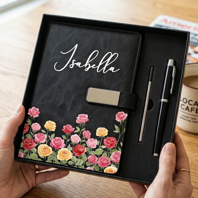 Puede incluir: Un juego de diario negro con un dise&ntilde;o floral de rosas y el nombre "Isabella" en escritura blanca. El juego incluye un bol&iacute;grafo y un l&aacute;piz &oacute;ptico, presentado en una caja de regalo negra. Las rosas son en tonos rosa, amarillo y rojo.