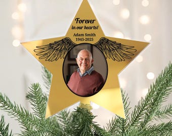 Può includere: Un ornamento commemorativo a forma di stella dorata con la foto di un uomo con un maglione bordeaux. L'ornamento presenta la scritta "Forever in our hearts!" e "Adam Smith 1945-2025" con decorazioni ad ali. L'ornamento è su un albero di Natale.