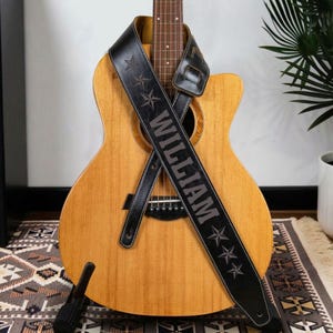 May include: A light brown acoustic guitar with a black leather guitar strap draped across the body. The strap features the name WILLIAM embossed in capital letters, flanked by three star icons on both the top and bottom sections of the strap.