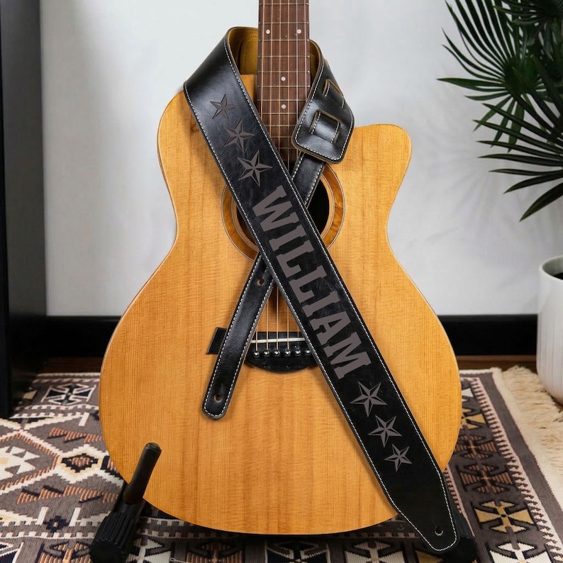 May include: A light brown acoustic guitar with a black leather guitar strap draped across the body. The strap features the name WILLIAM embossed in capital letters, flanked by three star icons on both the top and bottom sections of the strap.