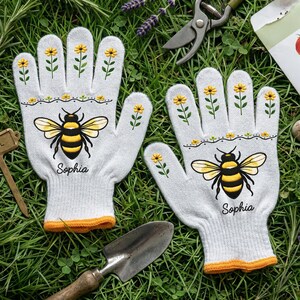 Op de afbeelding: Een paar witte tuinhandschoenen met een gele en zwarte bijenillustratie, kleine gele bloempatronen op de vingers en de naam Sophia in zwarte letters. De handschoenen hebben oranje geribbelde boorden en zijn gemaakt van gebreide stof.