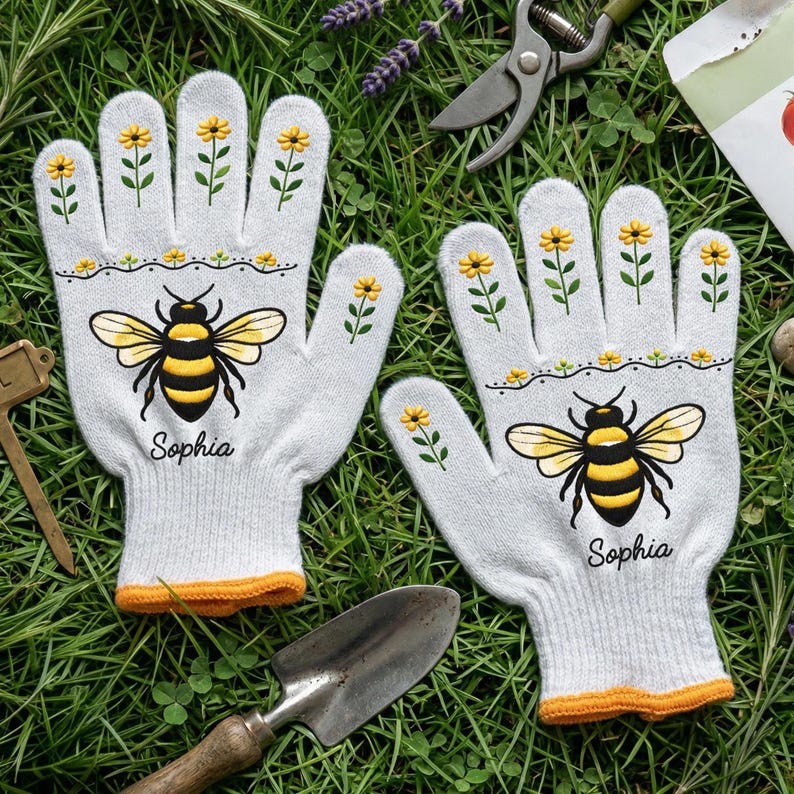 Op de afbeelding: Een paar witte tuinhandschoenen met een gele en zwarte bijenillustratie, kleine gele bloempatronen op de vingers en de naam Sophia in zwarte letters. De handschoenen hebben oranje geribbelde boorden en zijn gemaakt van gebreide stof.