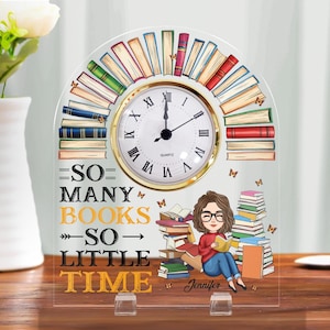 May include: A clear acrylic desk clock with a gold-rimmed clock face and a design featuring books and a person reading. The clock reads "SO MANY BOOKS SO LITTLE TIME" with the name "Jennifer" at the bottom. The clock is supported by two clear stands.