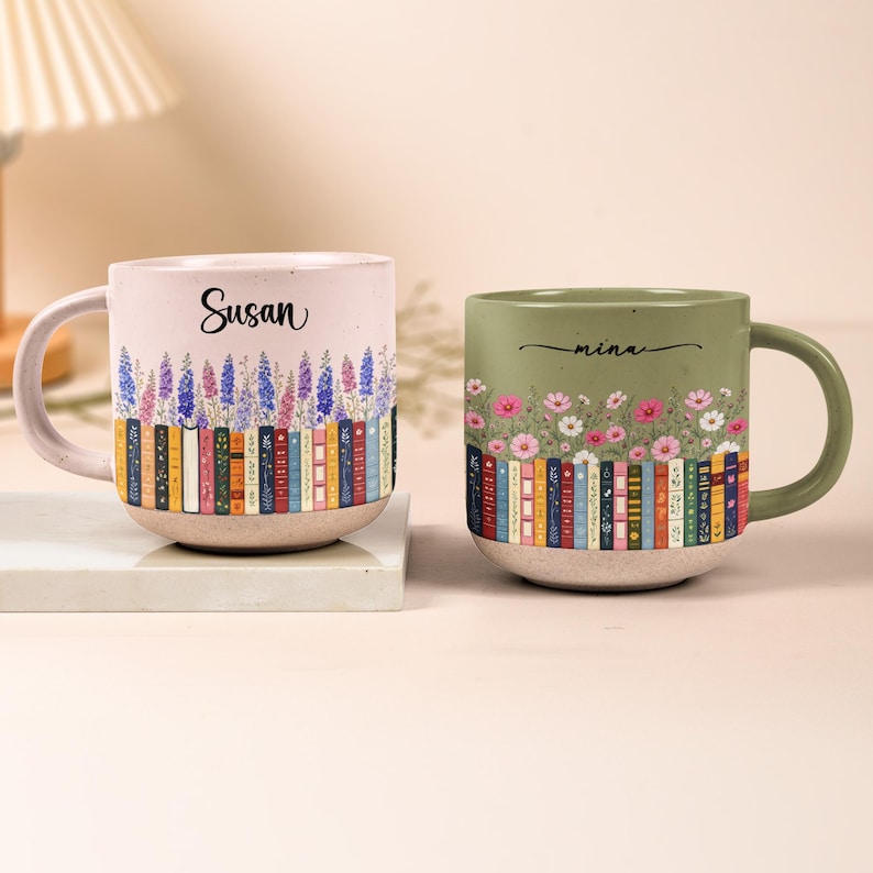 May include: Two ceramic mugs with speckled finishes. The left mug is light pink with the name Susan, purple and blue flowers, and a row of colorful books. The right mug is sage green with the name mina, pink and white flowers, and colorful books.