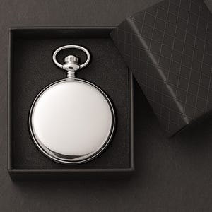 Puede incluir: Un reloj de bolsillo plateado con un acabado pulido se presenta en una caja negra. El reloj tiene una esfera circular y un lazo. La caja est&aacute; abierta, revelando una tapa texturizada.