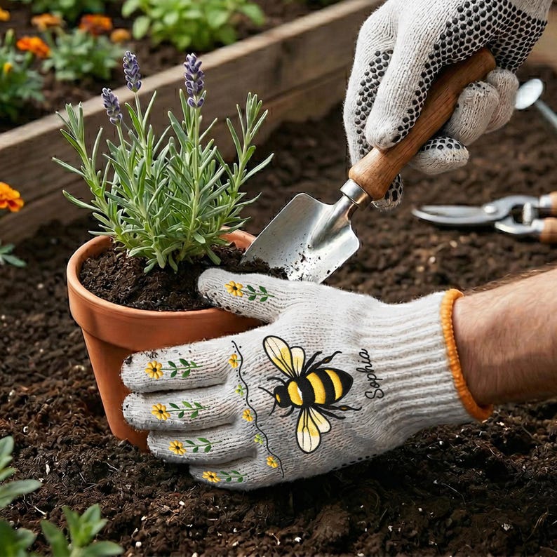 Op de afbeelding: Een paar lichtgrijze tuinhandschoenen met een gele bij-afbeelding, kleine gele bloempatronen en de naam Sophia erop gedrukt. De handschoenen hebben oranje boorden en zwarte gripstippen, getoond terwijl ze een terracotta pot met lavendel vasthouden.