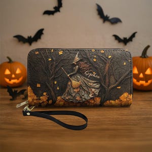 May include: A black wallet with a witch illustration, a broom, and the name "Sophia" in script. The wallet features a wrist strap and a zipper closure. The background includes fall leaves, trees, and Halloween decorations.