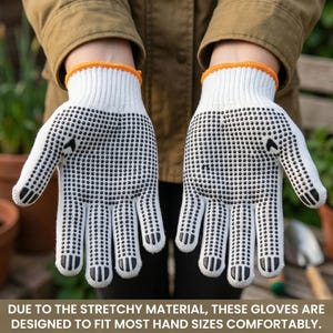 Op de afbeelding: Een paar witte gebreide handschoenen met zwarte stippenpatronen op de handpalmen en vingers, afgewerkt met een oranje elastische boord. De handschoenen hebben een gestructureerd oppervlak voor een betere grip tijdens het werken.