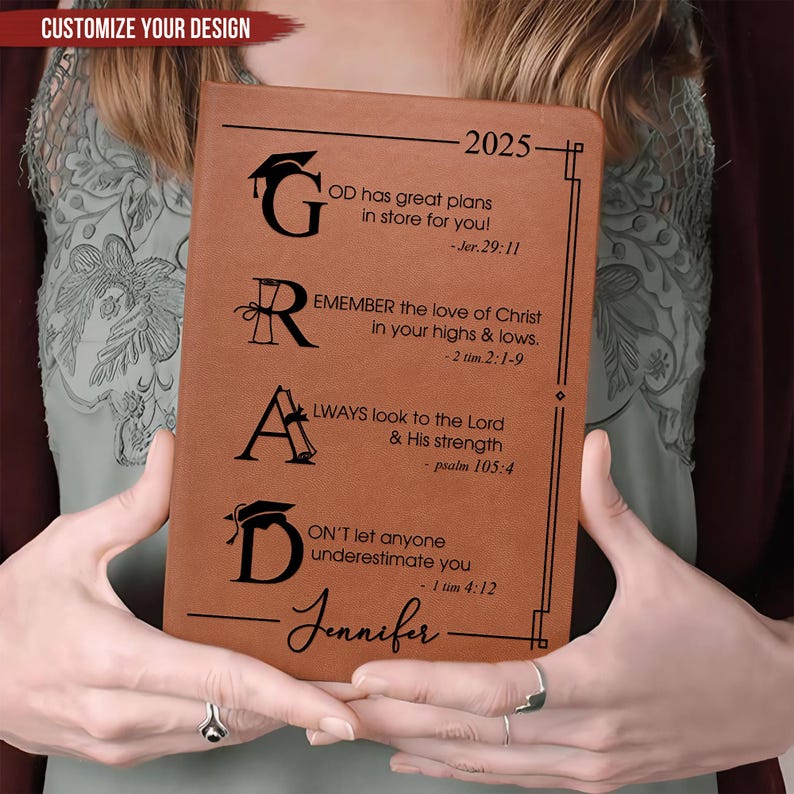 May include: A brown leather journal with the word "GRAD" in black lettering. The journal has inspirational quotes and verses from the Bible, including "God has great plans in store for you!" and "Don't let anyone underestimate you." The journal is personalized with the name "Jennifer" and the year "2025".