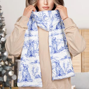 May include: White scarf with a blue toile pattern featuring dogs, trees, and landscapes. The scarf is wrapped around the neck and shoulders. The scarf is made of a soft, quilted material.
