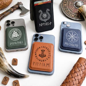 May include: A collection of phone cases with attached cardholders, featuring various Norse symbols and runes. The cases are in shades of green, black, blue, and brown. The image includes an axe, a shield, and wooden runes, creating a Viking theme.