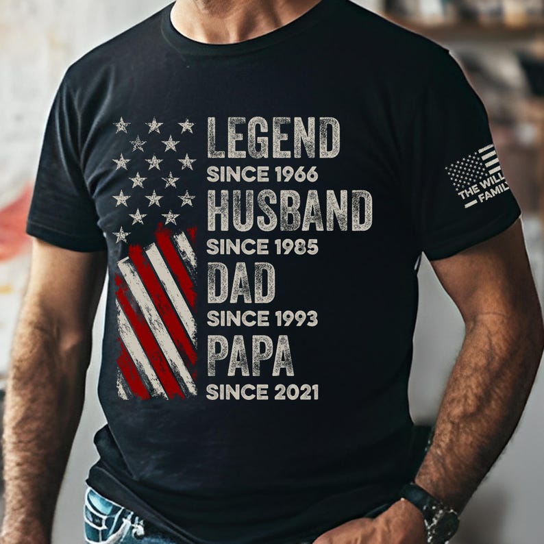 May include: Black t-shirt with a distressed American flag design. The shirt features the words "LEGEND SINCE 1966, HUSBAND SINCE 1985, DAD SINCE 1993, PAPA SINCE 2021" in white lettering. The text "THE WILL - FAMILY" is printed on the right sleeve.