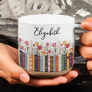 May include: A white ceramic mug with a speckled base featuring a colorful illustration of a row of books with various floral patterns and a field of tulips above them. The name Elizabeth is printed in black cursive script on the upper portion.