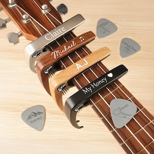 Könnte beinhalten: Akustikgitarre mit vier personalisierten Kapodastern und Plektren. Die Kapodaster sind silber, braun, helles Holz und schwarz, jeweils mit einem Namen oder Satz graviert: "Claire", "Michael", "A.J" und "My Honey". Gitarrenplektren sind mit Namen und Designs graviert.