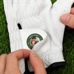 K&ouml;nnte beinhalten: Ein wei&szlig;er Golfhandschuh mit einem kreisf&ouml;rmigen Aufn&auml;her am verstellbaren Handgelenkriemen. Der Aufn&auml;her hat einen dunkelgr&uuml;nen Rand und zeigt ein Foto eines Mannes, der ein Kind h&auml;lt, mit dem wei&szlig;en Schriftzug BEST DAD am oberen Rand.