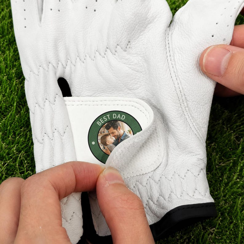 K&ouml;nnte beinhalten: Ein wei&szlig;er Golfhandschuh mit einem kreisf&ouml;rmigen Aufn&auml;her am verstellbaren Handgelenkriemen. Der Aufn&auml;her hat einen dunkelgr&uuml;nen Rand und zeigt ein Foto eines Mannes, der ein Kind h&auml;lt, mit dem wei&szlig;en Schriftzug BEST DAD am oberen Rand.