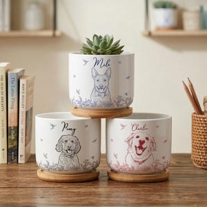 May include: Three white ceramic planters with dog portraits and names, each on a wooden saucer. The top planter has a succulent. The dogs are named Milo, Penny, and Charlie. Decorative birds and floral accents surround the dog portraits.
