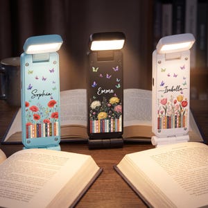 Könnte beinhalten: Drei tragbare LED-Buchleuchten in Hellblau, Schwarz und Weiß. Jede Leuchte hat einen rechteckigen Kopf, einen vertikalen Körper mit Blumen- und Buchrückenillustrationen sowie einen in Schreibschrift gedruckten Namen: Sophia, Emma und Isabella.