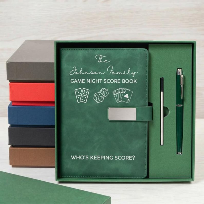 May include: A green game night score book with a silver metal clasp, featuring the text The Johnson Family GAME NIGHT SCORE BOOK and WHO'S KEEPING SCORE? along with icons of dice and playing cards, presented in a box with a matching green pen.