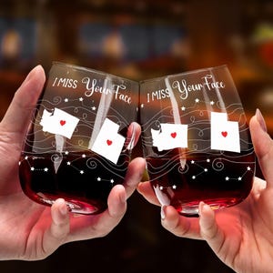 May include: Two stemless wine glasses with the text "I Miss Your Face" and outlines of the states of Washington and Utah. The glasses are filled with red wine and are being clinked together.