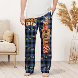 May include: Blue plaid pajama pants with the text "COOKIE CREW" and the name "JAMES" in gingerbread cookie lettering. The pants are decorated with colorful string lights.