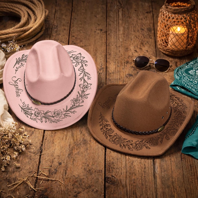 Pode incluir: Um chap&eacute;u de cowboy rosa claro com estampa floral preta e o nome Sophia, e um chap&eacute;u de cowboy marrom com estampa floral preta e o nome Emily. Ambos os chap&eacute;us possuem uma faixa tran&ccedil;ada escura com um pequeno detalhe met&aacute;lico na frente.