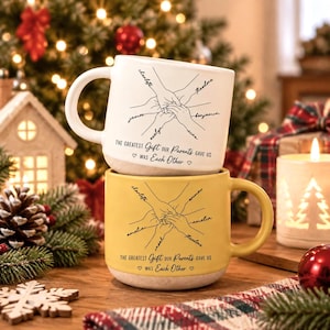 May include: Two ceramic mugs stacked vertically, one white and one yellow, each featuring a black line drawing of multiple hands joined together in the centre. The text reads: The greatest gift our parents gave us was each other, with names written along the arms.