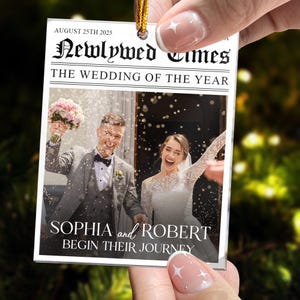 May include: A rectangular hanging ornament designed like a newspaper front page titled Newlywed Times. It features a photo of a couple in wedding attire with confetti, the date August 25th 2025, and the text Sophia and Robert Begin Their Journey.