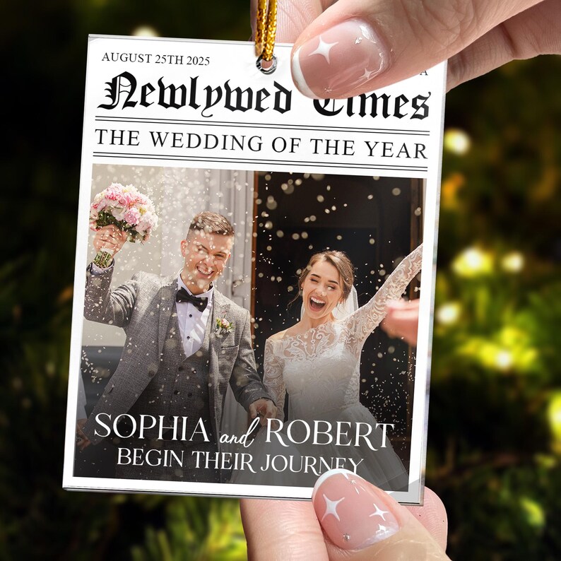 May include: A rectangular hanging ornament designed like a newspaper front page titled Newlywed Times. It features a photo of a couple in wedding attire with confetti, the date August 25th 2025, and the text Sophia and Robert Begin Their Journey.