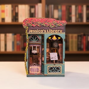 May include: A miniature wooden model of "Jessica's Library" with a detailed facade. The model features a teal and brown color scheme, a floral roof, and a clock. The front door has an "Open" sign. The model is set against a blurred background of bookshelves.