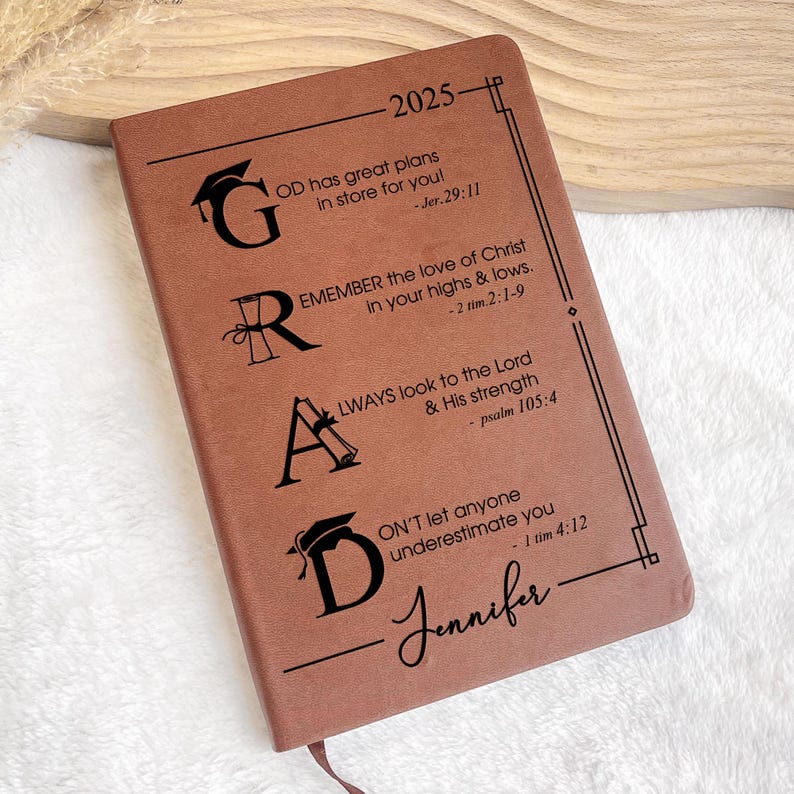 May include: A brown leather journal with the word "GRAD" in black lettering. Each letter is decorated with a graduation cap. The journal has inspirational quotes and bible verses about faith and perseverance. The year 2025 and the name "Jennifer" are also printed on the cover.