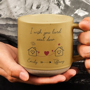 May include: A yellow ceramic mug with a brown bottom. The mug has a white inscription that reads "I wish you lived next door" with two houses and hearts connected by a line. The names "Emily" and "Tiffany" are written below the houses.