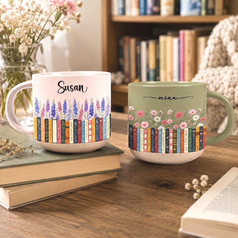 May include: Two ceramic mugs featuring a row of colorful book spines at the base topped with floral illustrations. The left mug is light pink with the name Susan, while the right mug is sage green with the name mina printed in black script.