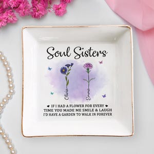 Può includere: Un vassoio portaoggetti in ceramica bianca con bordo dorato che presenta un design ad acquerello di due fiori con il testo "Soul Sisters" e i nomi "Olivia" e "Emma" sotto i fiori. Il testo sotto i fiori dice "Se avessi un fiore per ogni volta che mi hai fatto sorridere e ridere, avrei un giardino in cui passeggiare per sempre."