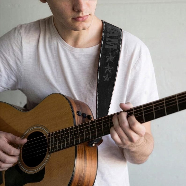 May include: A brown acoustic guitar with a black guitar strap featuring a vertical row of three grey stars and the letters AM. The strap has white stitching along the edges and is worn over a plain white t-shirt. The guitar has a dark pickguard.