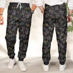 May include: Black sweatpants with a repeating pattern of neon-colored video game controllers, lightning bolts, and the name "TOM". The pants have a drawstring waist and elastic cuffs. White sneakers complete the look.