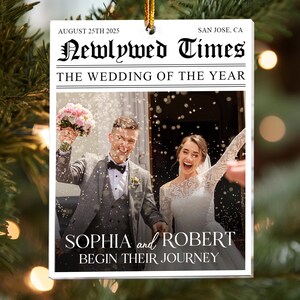 May include: A rectangular white ornament featuring a newspaper-style design titled Newlywed Times. It includes the text August 25th 2025, San Jose, CA, The Wedding of the Year, and Sophia and Robert Begin Their Journey, with a photo of a couple.