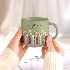 May include: A sage green ceramic mug featuring a printed design of a row of colorful books topped with white lily flowers. The name Sophia is written in black cursive script above the floral illustration. The mug has a smooth handle and a base.