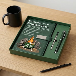 May include: A dark green gift set containing a notebook with a silver clasp, a pen, and a matching mug. The notebook cover features the text Dumpster Fire Response Team and an illustration of raccoons around a burning dumpster with office items.