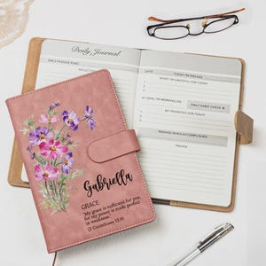 May include: A dusty rose daily journal with a floral print, the name Gabriella, and the text Grace My grace is sufficient for you, for my power is made perfect in weakness. 2 Corinthians 12:9. Includes an open page with daily prompts.