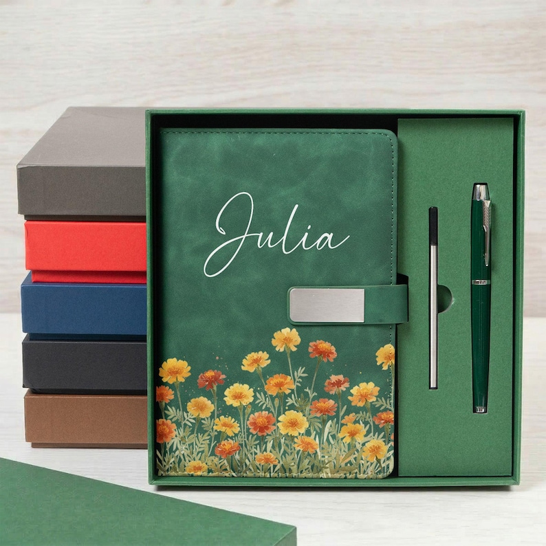 Puede incluir: Un juego de diario y bol&iacute;grafo verde con el nombre "Julia" en escritura blanca. El diario presenta un dise&ntilde;o floral con flores amarillas y naranjas. El juego se presenta en una caja de regalo verde, con una pila de cajas coloridas en el fondo.