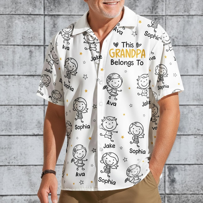 Peut inclure: Chemise blanche &agrave; boutons avec un motif r&eacute;p&eacute;t&eacute; de dessins d'enfants et d'&eacute;toiles. Le texte "This Grandpa Belongs To" en or est pr&eacute;sent, avec des noms comme Ava, Jake et Sophia.