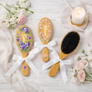 May include: Three wooden hairbrushes with white ribbon bows. Two brushes are decorated with floral designs and names, one with purple violets and the name "Sophia", the other with pink flowers and the name "Miranda". The third brush has black bristles.