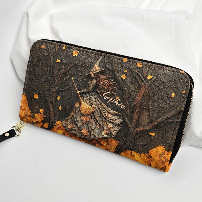 May include: A zippered wallet featuring a witch in a forest scene with autumn leaves. The witch is holding a broom and wearing a hat and dress. The name "Sophia" is written on the wallet. The wallet has a black zipper and a gold-colored wrist strap.