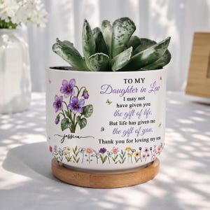 May include: A white ceramic planter with a bamboo saucer containing a green succulent. The planter features a purple violet illustration, the name Jessica, and text reading: To my Daughter in Law, I may not have given you the gift of life.