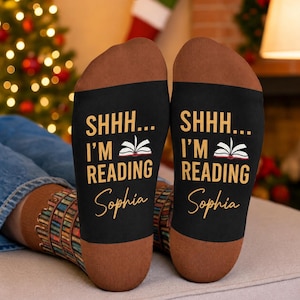 May include: A pair of black socks with brown heels and toes. The text SHHH... I'M READING is printed in yellow above a white open book icon, followed by the name Sophia in yellow script. The lower leg section features a colourful book spine pattern.