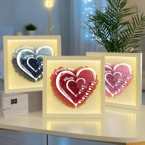 May include: Three illuminated heart-shaped wall art pieces. Each features a heart design made of paper roses, with the words "Grandma" and "Mom" and names. The frames are white, and the roses are in shades of blue, red, and pink.